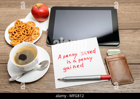 Tovagliolo scrittura proverbio del messaggio sul tavolo di legno con caffè, alcuni alimenti e tablet PC un penny risparmiato è un penny guadagnato Foto Stock