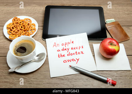 Tovagliolo scrittura proverbio del messaggio sul tavolo di legno con caffè, alcuni alimenti e tablet PC Una mela al giorno toglie il medico di torno Foto Stock