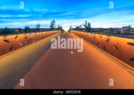 Illuminato dallo stile moderno decorativo creazione di rusty ponte per pedoni e biciclette oltre l'acqua durante il crepuscolo in una città suburbane. Case residenziali su t Foto Stock