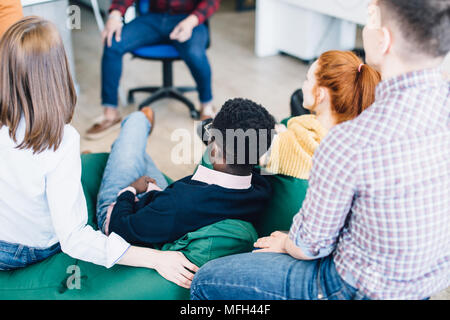 Vista dal lato posteriore del giovane diversi studenti seduti sul verde pouf incentrato su coach training. giovani dipendenti in sede di riunione Foto Stock