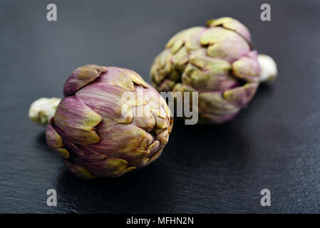 Due carciofi freschi con stelo su sfondo di ardesia closeup Foto Stock