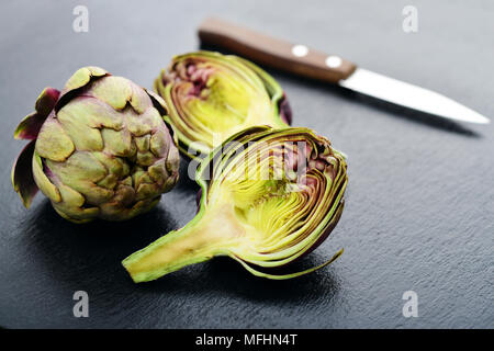 Due carciofi freschi con stelo e una metà che mostra il cuore su sfondo di ardesia closeup Foto Stock