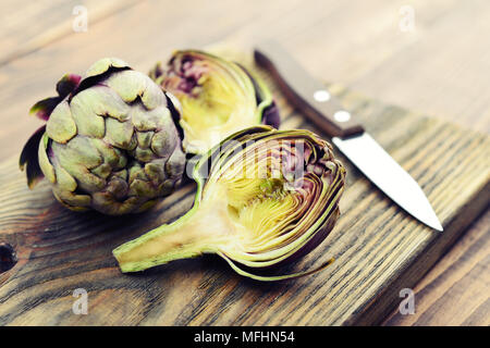 Due carciofi freschi con stelo e una metà che mostra il cuore su sfondo di legno closeup Foto Stock