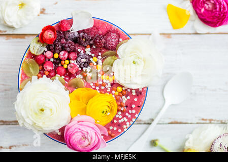 La colazione Detox frullato rosa da banana e frutti di bosco nella ciotola, guarnita con frutti di bosco surgelati e ranunculus fiori, luce sullo sfondo di legno, Foto Stock