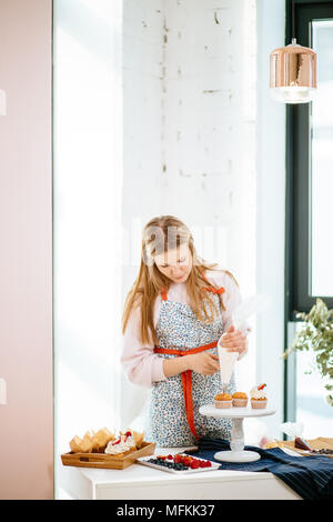 A mezza lunghezza Ritratto di pasticceria blondy femmina nel bel grembiule, la decorazione di pane appena sfornato tortine con glassa cremosa Foto Stock