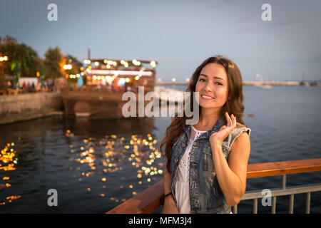 Piuttosto giovane donna camminare sul lungomare della città vicino al mare di sera. Colori tenui. Le luci di strada raggi cade sul suo viso. Felice ragazza gode di vacanza Foto Stock