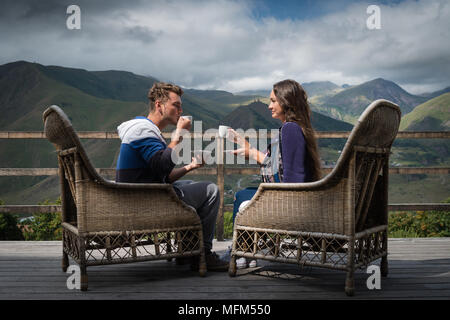 Coppia giovane di buon umore si siede sulla terrazza con tazze di caffè al mattino con bella vista. Bel ragazzo con la fidanzata avente la prima colazione. Foto Stock