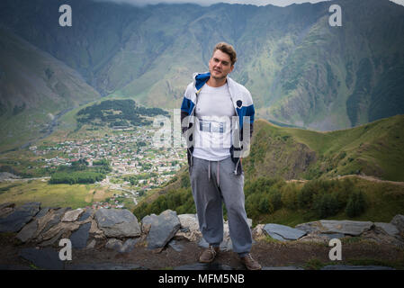 Giovane uomo sorge sulla sommità e sorrisi. Traveler in abiti casual su sfondo di piccola città nella valle e le maestose montagne. Foto Stock