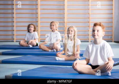 Concentrati gli alunni seduti con le gambe incrociate su tappeti di blu durante la classe di ginnastica Foto Stock