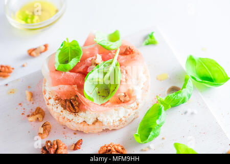 Pane croccante snack sani con prosciutto di Parma, soft cottage cheese, noci, olio d'oliva, paprica, di foglie di basilico fresco. Facile colazione close-up su una bianca bac Foto Stock