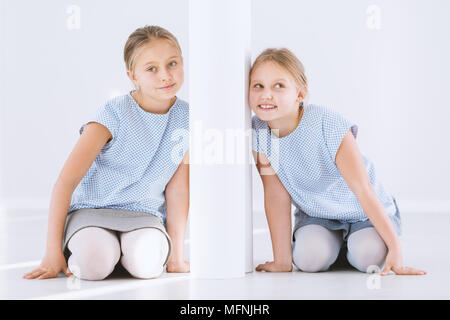 Ritratto di giovane, felice gemelle in ginocchio e appoggiata contro i lati opposti di un muro bianco Foto Stock