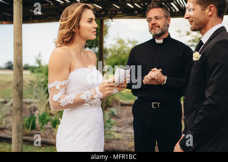 Bella giovane sposa che legge le promesse nuziali da un giornale. rituali di cerimonia nuziale di giovani coppie. Foto Stock