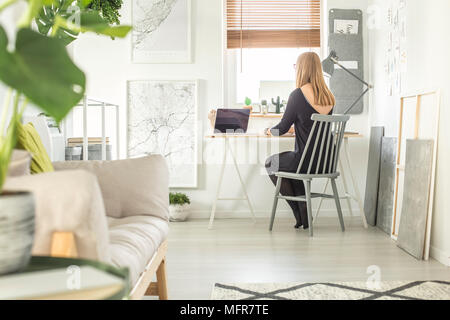 Ragazza seduta alla scrivania che lavora su un computer in un ufficio a casa interno con un divano, un poster e un muro grigio organizzatore