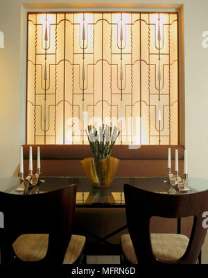 Un dettaglio di una moderna sala da pranzo con tavolo in vetro sedie poltrone panchetto parete decorativa grill Foto Stock