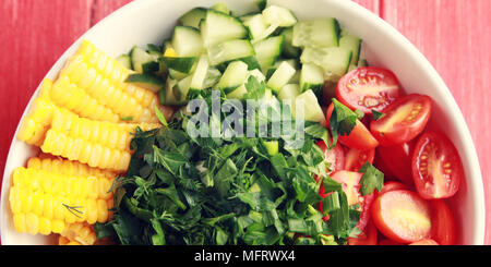 Piastra bianca con verdure tagliate per un insalata vegana in rosso di un tavolo di legno. Foto colorate. Il mais, pomodori ciliegini, il peperone, la cipolla, il cetriolo e gar Foto Stock