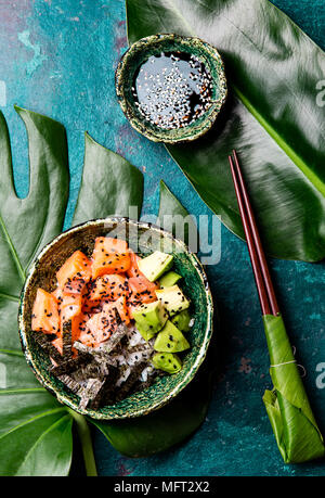 Hawaiian poce di salmone con avocado, riso e sesamo servito in ciotole su foglie tropicali. Turchese sullo sfondo di ardesia. Vista dall'alto. Foto Stock