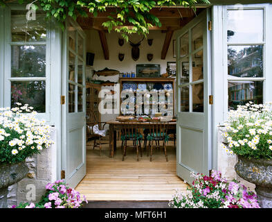 Fiori che fioriscono intorno a porte francesi si aprono in un paese zona pranzo con tavolo da pranzo e sedie, pavimento in legno e una collezione di stoviglie su open shel Foto Stock