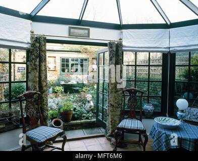 Conservatorio dettaglio con un tetto di vetro, tende, tavolo rotondo e sedie che fiancheggiano aprire porte francesi all' interno del giardino. Foto Stock