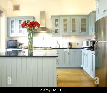 Moderna cucina di paese con verde verniciato, vetro anteriore di armadi, isola centrale e pavimento in legno. Foto Stock