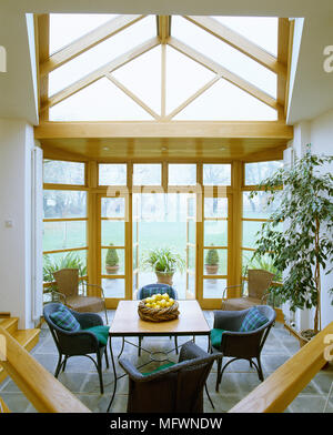 Una moderna serra con un tetto di vetro del tetto, tavolo da pranzo con sedie di vimini e una vista attraverso aprire porte francesi ad un prato erboso. Foto Stock