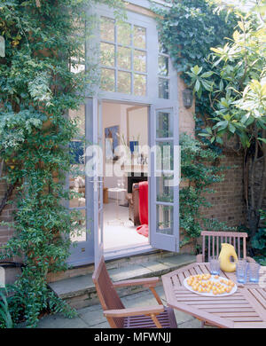 Una vista esterna della casa francese che mostra le porte aperte a un patio pavimentato area, tavolo in legno e sedie, piante rampicanti Foto Stock