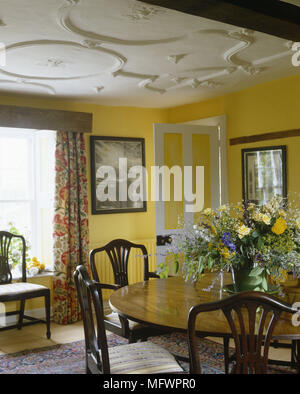 Circolare di tavolo da pranzo in legno con fiori e sedie in camera con pareti gialle e gesso dettaglio sul soffitto Foto Stock