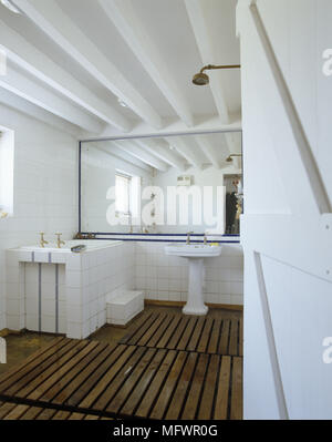 Bagno con pavimentazione in legno e pareti piastrellate di bianco con specchio vicino al lavandino e vasca da bagno Foto Stock