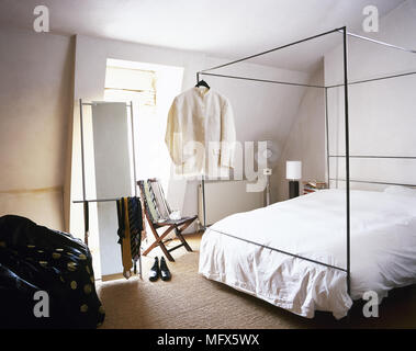 Camera da letto in stile contemporaneo con pavimento in moquette, soffitto inclinato, soleggiata finestra e uomini vestiti appesi da un metallo letto a baldacchino. Foto Stock