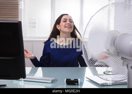 Sorridente giovane imprenditrice stessa di raffreddamento con ventola elettrica durante la stagione calda Foto Stock