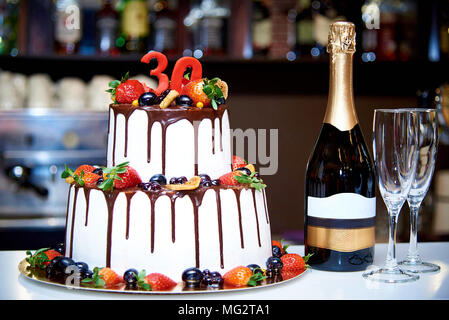 Un due-tiered white torta con frutta fresca e cioccolato sorge accanto ad una bottiglia di champagne e due bokalam sullo sfondo di una barra sfocate in un re Foto Stock