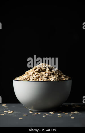 Primo piano di un bianco di ceramica ciotola piena di fiocchi d'avena su una tabella grigia, contro uno sfondo nero con un po' di spazio vuoto sulla parte superiore Foto Stock