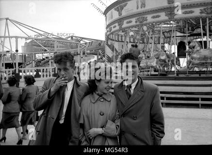 3 ben vestito di moda teenager, 2 ragazzi e una ragazza, in una fredda giornata presso un parco di divertimenti britannico negli anni cinquanta, si sta fumando una sigaretta. Foto Stock