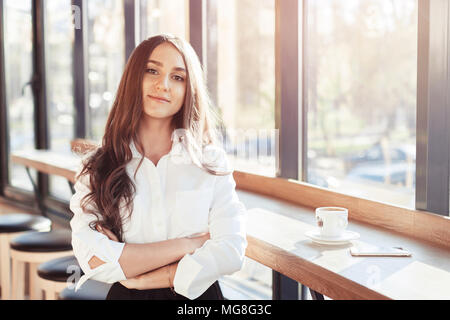 Ragazza di business in camicia bianca in cafe sorridente e guardando la fotocamera. Una ragazza beve caffè in una rottura tra il lavoro. La ragazza lavora in un caffè Foto Stock