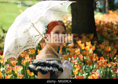 Una donna con i capelli rossi e un ombrellone bianco per proteggersi dal sole si rilassa in un campo di rosso e tulipani gialli Foto Stock