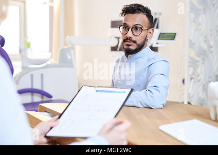 Dentista femmina indossa mantello bianco seduto alla scrivania in ufficio e il riempimento in medical record durante la consultazione di mixed-gara paziente giovane Foto Stock