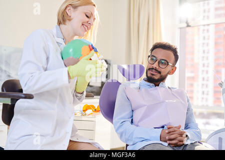 Sorridente giovane dentista indossando guanti di gomma e mantello bianco utilizzando il modello anatomico del cranio umano mentre per spiegare il metodo di trattamento per il paziente barbuto Foto Stock