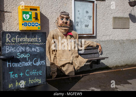Il vecchio uomo spaventapasseri seduto sul sedile accanto al defibrillatore di emergenza box in Wray, Lancaster, Regno Unito. 28/04/2018. Wray Spaventapasseri Festival e village cafe.gli abitanti del villaggio sono tornati di nuovo per la creazione di alcuni strani e bizzarri e meravigliosi spaventapasseri le creazioni di sorprendere e deliziare il visitatore. Ogni anno i visitatori girovagare Wray village, scoprendo l'host di scarecrows che hanno schioccato in su nei giardini del villaggio. Credito: MediaWorldImages/AlamyLiveNews Foto Stock