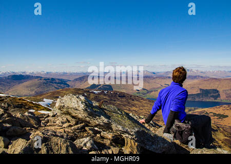 Un maschio di escursionista si siede, appoggia e guarda a distanza in una limpida giornata di sole alla vista delle Highlands Scozzesi dal vertice del Ben più, Trossachs Foto Stock