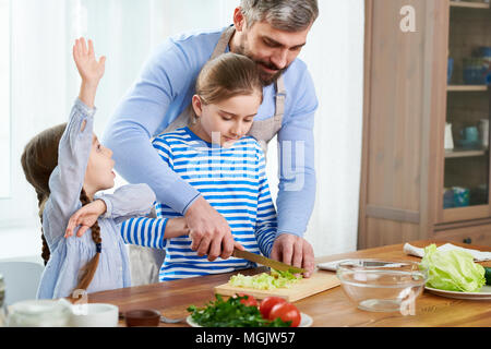 Uomo bello indossare grembiuli ad insegnare la sua piccola figlia di cavolo tagliato correttamente mentre avvolto in preparazione di insalata di verdure, altra figlia con ra Foto Stock