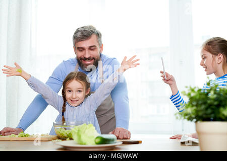 Vista di profilo di sorridente bambina di scattare una foto del suo bel papà e carino sorella mentre essi avvolto in preparazione di insalata di verdure, cucina int Foto Stock