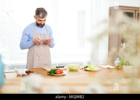 Barbuto di mezza età food blogger che indossa il grembiule permanente al tavolo di legno e fotografare gli ingredienti per la deliziosa insalata di verdure su tavoletta digitale Foto Stock