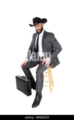 Un bel giovane uomo d affari che indossa un vestito e un cappello da cowboy seduta su una poltrona con la sua valigetta, isolato per sfondo bianco Foto Stock