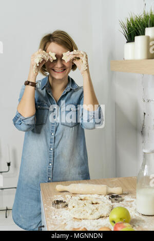 Giovane ragazza attraente è la preparazione di biscotti in cucina. Essa contiene pezzi di pasta nelle sue mani. Ragazza stolti intorno a chiudere gli occhi. Latte, mele, farina Foto Stock