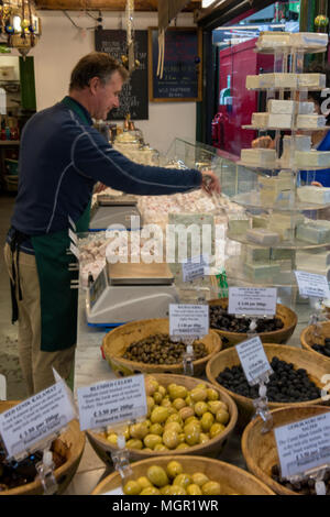 Un titolare di stallo o commerciante di mercato la vendita di olive fresche e specialità continentali formaggi in una fase di stallo sul mercato di Borough londinese di alta qualità per alimenti. Foto Stock