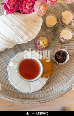 Carcade tè in una tazza bianca con i cookie sul rustico vassoio di metallo e con ranuncoli rosa in un vaso e tre candele beige Foto Stock