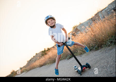 Sorridente ragazzino nello sport equitazione casco su scooter sulla strada abbandonati in estate caldo tramonto Foto Stock