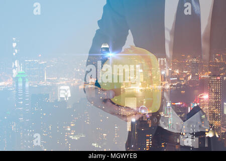 La doppia esposizione concetto di ingegnere con sicurezza Casco giallo per la sicurezza dei lavoratori con la città moderna notte background. concetto di settore. Foto Stock
