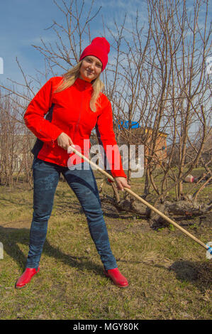 Giovane bella donna agricoltore pulisce rastrello erba secca, indossa una giacca rossa, stivali e cappello, foto verticale Foto Stock