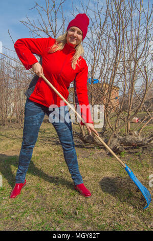 Giovane bella donna agricoltore pulisce rastrello erba secca, indossa una giacca rossa, stivali e cappello, foto verticale Foto Stock