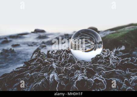 Sfera di vetro sulle foglie congelate in inverno in natura ruvida con colline Foto Stock